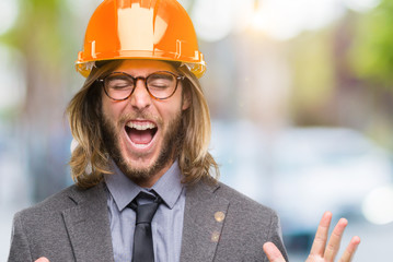 Sticker - Young handsome architec man with long hair wearing safety helmet over isolated background celebrating mad and crazy for success with arms raised and closed eyes screaming excited. Winner concept