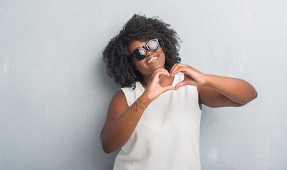 Poster - Young african american plus size woman over grey grunge wall wearing fashion sunglasses smiling in love showing heart symbol and shape with hands. Romantic concept.