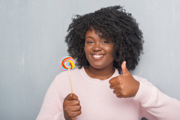 Sticker - Young african american woman over grey grunge wall eating lollipop candy happy with big smile doing ok sign, thumb up with fingers, excellent sign