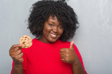 Wall Mural - Young african american woman over grey grunge wall eating chocolate chip cooky happy with big smile doing ok sign, thumb up with fingers, excellent sign