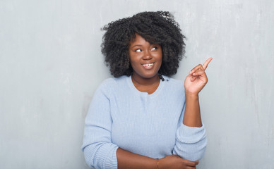 Wall Mural - Young african american woman over grey grunge wall wearing winter sweater with a big smile on face, pointing with hand and finger to the side looking at the camera.