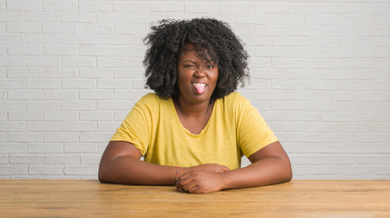 Poster - Young african american woman sitting on the table at home sticking tongue out happy with funny expression. Emotion concept.