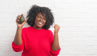 Canvas Print - Young african american woman over white brick wall eating avocado screaming proud and celebrating victory and success very excited, cheering emotion