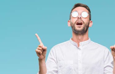 Young handsome man wearing glasses over isolated background amazed and surprised looking up and pointing with fingers and raised arms.