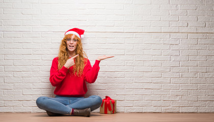 Wall Mural - Young redhead woman sitting over brick wall wearing christmas hat amazed and smiling to the camera while presenting with hand and pointing with finger.