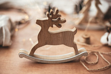 Merry Christmas. Rustic Reindeer Christmas Toy On Wooden Table On Background Of Lights, Wooden Tree, Twine, Gift In Linen With Green Branch, Pine Cones. Space For Text. Simple Eco Presents.