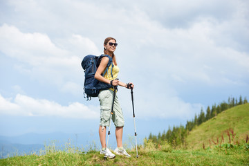 attractive smiling woman tourist hiking mountain trail, walking on grassy hill, wearing backpack and
