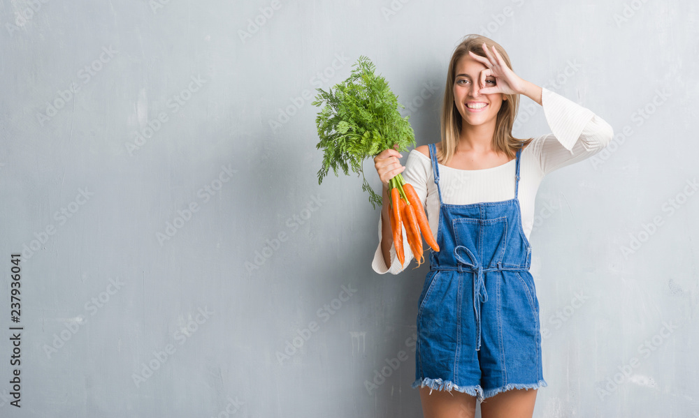 Beautiful young woman over grunge grey wall holding fresh carrots with happy face smiling doing ok sign with hand on eye looking through fingers - obrazy, fototapety, plakaty 