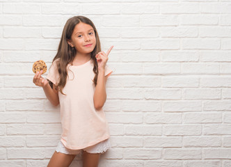 Young hispanic kid over white brick wall eating chocolate chips cooky very happy pointing with hand and finger to the side