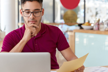 Man professional deliberates business idea, works on writing new article, checks information from paper and laptop device, reads interesting electronic book, wears eyeglasses, keeps hand on chin