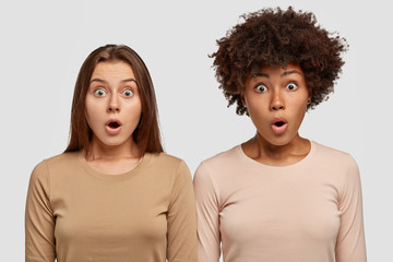 Wall Mural - Stupefied mixed race two young women have bated breath, opens mouth with surprisement, dressed in casual clothes, stand shoulder to shoulder, isolated over white background. Reaction concept