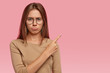 © WHstudio Leushin N - Displeased European woman with dark hair, purses lower lip, has dejected facial expression, points aside with index finger, dressed in casual clothes, isolated over pink wall, dislikes item.