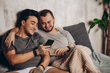 Wall Mural - Cheerful same-sex couple relaxing in bedroom, looking at phone with excitement and smiling joyfully remembering sweet moments of last night party