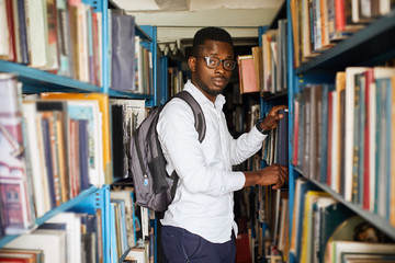 Wall Mural - Portrait of african university student with backpack over his shoulders visiting library in search of proper information and looking at camera. Horizontal shape, waist up, front view