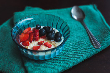 High angle view on cooked oat with forest fruits. Strawberries and blueberries in a blue glass bowl on a green napkin.