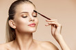 Beautiful woman using brush for brows. Photo of attractive woman with perfect makeup on beige background. Beauty concept
