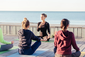 Group of healthy sporty young people resting after an outdoor yoga group training with attractive female Instructor of teacher, breathing deep. Yoga or pilates practice by the sea