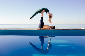 Acro yoga concept. Pair yoga. Couple of young sporty people practicing yoga lesson with partner, man and woman in yogi exercise, arm balance pose, working out by pool, above beach, against blue sky