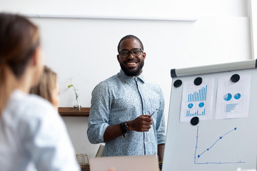 Black coach presents for company members financial report standing near whiteboard where shown sales statistics in graphs. Confident company owner teach interns acquaints with nuances of the market