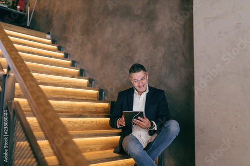 Photographie Smiling middle-aged Caucasian businessman dressed smart casual sitting on the stairs in cafe while using tablet for video call