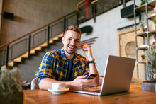 Portrait of young smiling Caucasian freelancer holding smart phone and putting on earphones while sitting in cafe. On the desk laptop and coffee.
