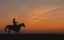 Cowboy In Hat Riding Horse On Colorful Cloudy Sky At Sunset. Silhouette Of  Cowboy Travel In Wild West Mountain Like Western Film Background