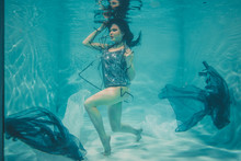Beautiful Model Girl Swims Under Water In A T-shirt In Gray Sequins And A Transparent Fashionable PVC Raincoat