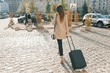 Young beautiful woman walking along city street with travel suitcase and cell phone. Fashionable brunette girl, view from the back, copy space
