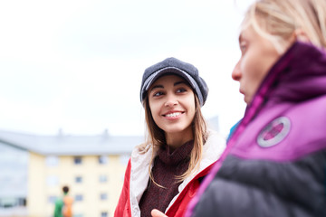 Wall Mural - Portrait of a young smiling woman in red jacket,gray scarf and cap. Woman walking, talking and laughing outdoors on the street with girlfriend at sunny autumn or spring day