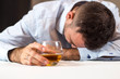 Pijany mężczyzna śpi przy stole z głową opartą o stół. W ręce trzyma szklankę whisky.