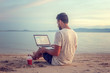 © olezzo - Young attractive guy a man with a beard sitting on the beach with a laptop on the background of the sea and sunset, distant work, freelancer, blogger, vacation and travel