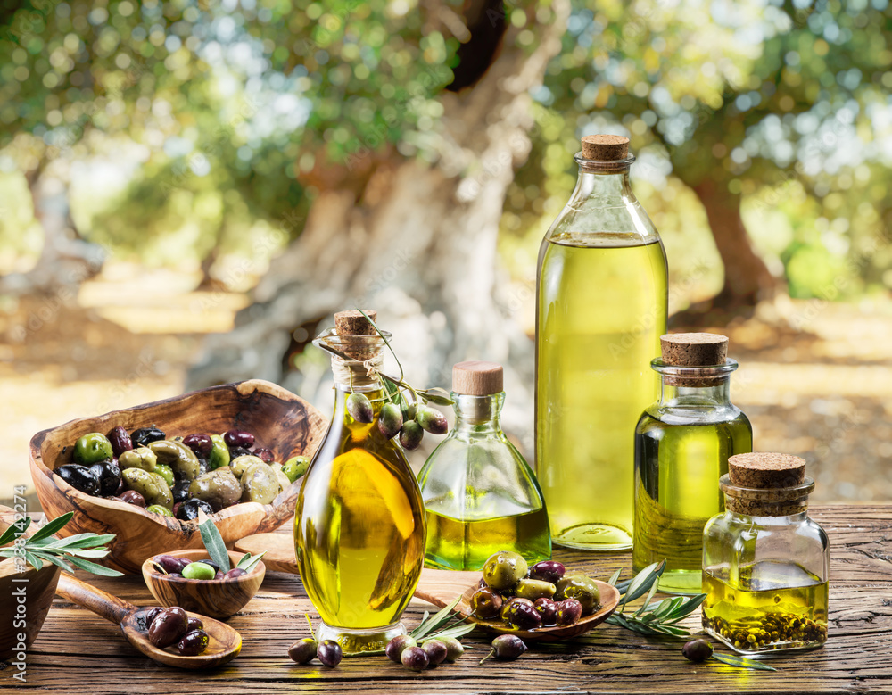 Olive oil and berries are on the wooden table under the olive tree. - obrazy, fototapety, plakaty 
