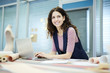 © pressmaster - Smiling attractive creative designer with curly hair leaning on table and looking away while using laptop in tailoring studio