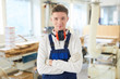 Content confident handsome young carpenter in ear protectors on neck wearing gloves and overall standing in woodworking shop and looking at camera