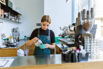 Experienced female barman making cappuccino. Woman barista in apron pouring hot milk foam into coffee. Latte art, small business and professional coffee brewing concept.