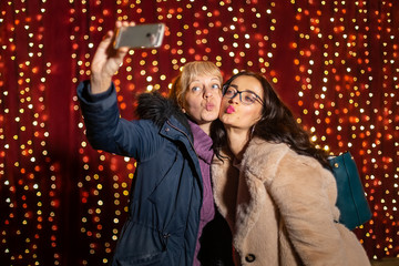 Wall Mural - Two attractive women taking selfie in front of light wall at Christmas market.