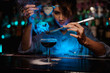 Female bartender pouring on the brown cocktail and on a flamed badian on tweezers a powdered sugar in the blue light