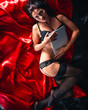 © Vitte Yevhen - Sexy young brunette woman in black sensual lingerie and in stockings using laptop on red bed in studio.