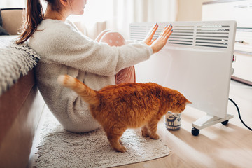 Using heater at home in winter. Woman warming her hands with cat. Heating season.