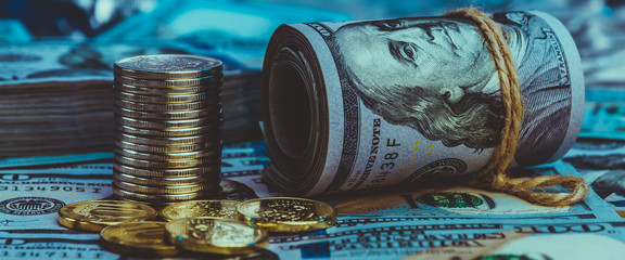 A roll of dollars with coins on the background of scattered one hundred dollar bills in blue light.