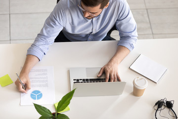 Wall Mural - Serious employee using laptop, typing, making notes in report with statistics and analysis at workplace, desk of office worker doing online job, development apps software, focused on project, top view