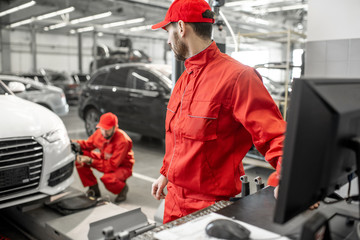 Two handsome auto mechanics in red uniform making wheel alignment with professional tools and computer at the car service