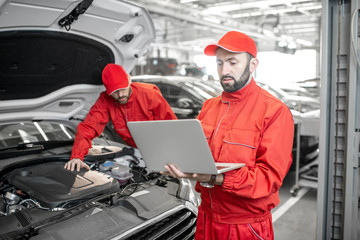 Two auto mechanics in red uniform doing engine diagnostics with computer in the car service