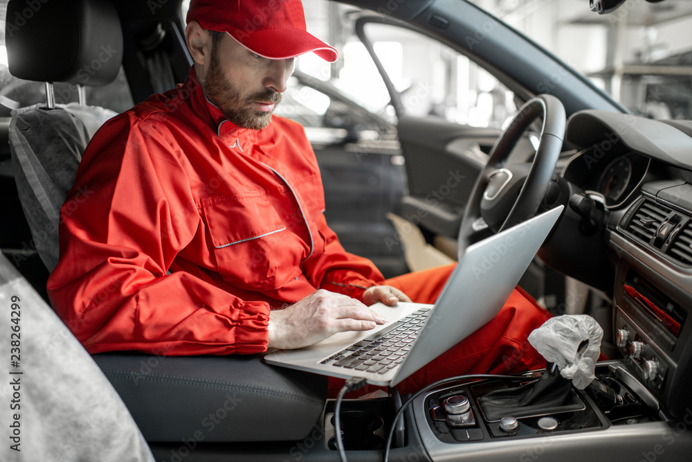 Auto mechanic in red uniform diagnosing car with computer sitting on ...