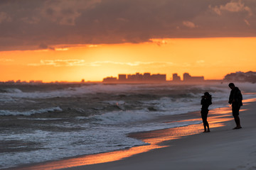 Wall Mural - Young couple looking at dramatic magical orange red sunset in Santa Rosa Beach, Florida with Pensacola coastline coast skyline in panhandle with ocean gulf mexico waves