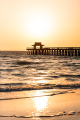 Wall Mural - Naples, Florida yellow and orange sunset in gulf of Mexico with sun setting by Pier wooden jetty, with horizon and dark silhouette ocean waves, sunlight reflection