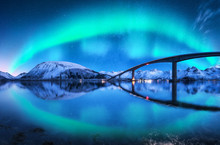 Bridge And Aurora Borealis Over Snowy Mountains. Lofoten Islands, Norway. Amazing Northern Lights And Reflection In Water. Winter Landscape With Starry Sky, Polar Lights, Road, Sea, City Illumination
