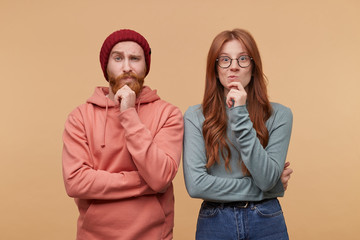 Wall Mural - Indoor shot of thoughtful male and female, holding chins and being concentrated on their problems. Young ginger couple dressed casualy thinking and have serious facial expression.