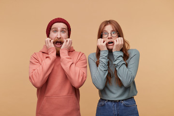 Wall Mural - horizontal shot of terrified young man and woman watch horror film together, keep hands near mouth, isolated over biege background