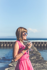 Wall Mural - Young woman in a long red dress and with a glass of wine posing on the sea background.